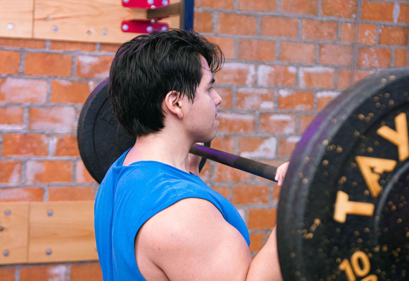 A man in a blue t-shirt holding a barbell across his shoulders in a brick-walled gym